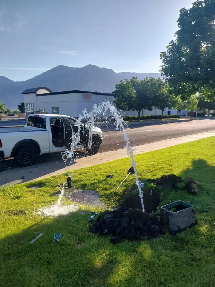 Water spraying in an arc from an open hole in a lawn beside a sidewalk, with mountains in the background